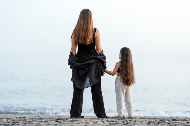 Beautiful young mother and little daughter holding hands and looking at the ocean at the beach