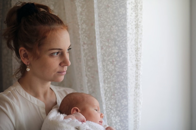 Beautiful young mother and newborn baby in her arms standing by the window