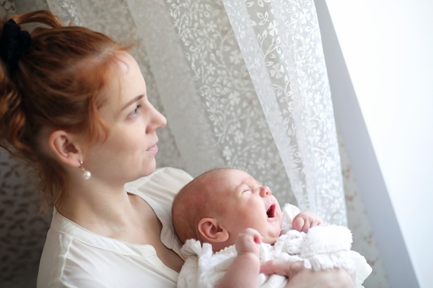 窓際に立っている彼女の腕の中で美しい若い母親と新生児