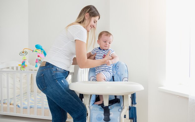 Beautiful young mother seating her baby boy in high chair