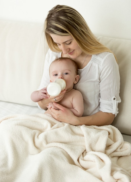 ベッドに座って哺乳瓶から赤ちゃんを養う美しい若い母親