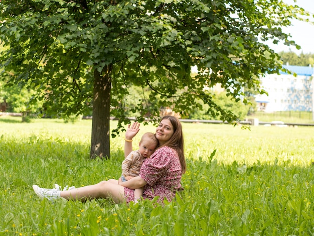 Beautiful young mother with a baby in her arms sits in the park on the grass on a sunny day