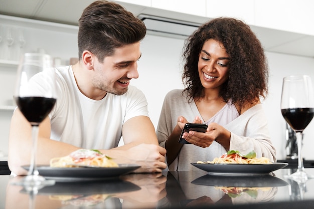 携帯電話を使用して、家でロマンチックな夕食を食べ、赤ワインを飲み、パスタを食べる美しい若い多民族のカップル