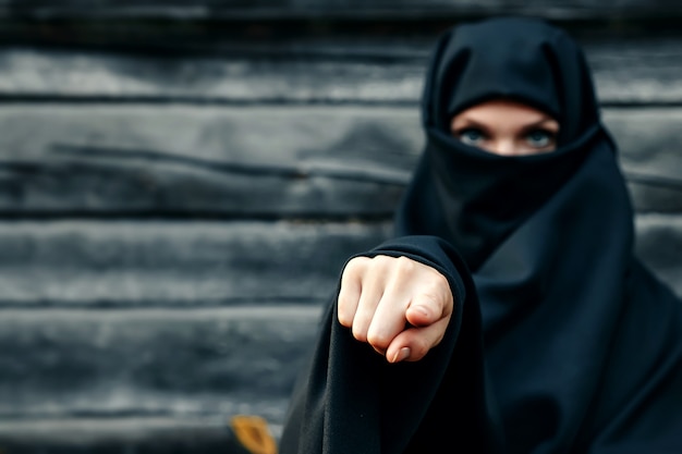 A beautiful, young, Muslim girl in a black veil with a closed face against a gray tree  points her finger at  camera. copyspace.