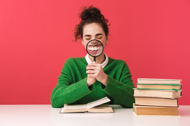 Beautiful young nerd student girl sitting at the table isolated, studying with books, holding magnifying glass