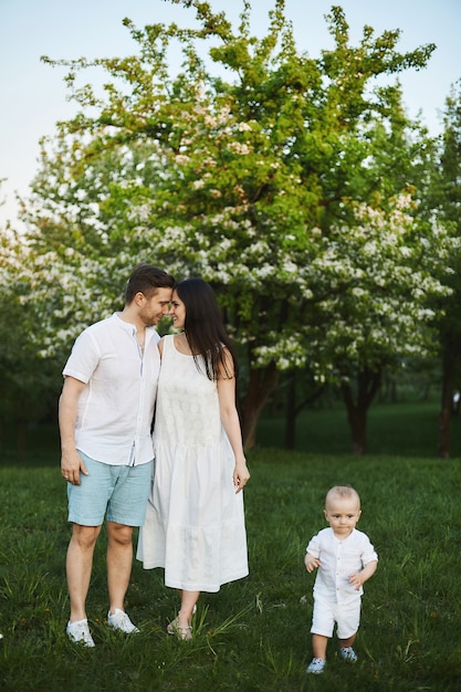 Beautiful young parents, stylish handsome man in shorts and shirt and brunette beauty in the white dress, and their cute little son have fun near trees at blooming park. Concept of a happy family