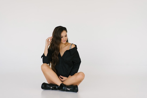 A beautiful young pregnant stylish girl in a black shirt and black shoes sits on the floor on a white  , she holds her hand on her stomach and dreams of a future baby