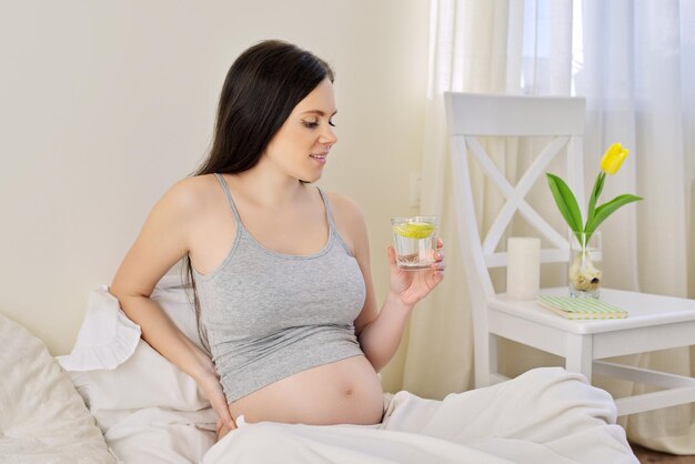 Beautiful young pregnant woman drinking water with lemon sitting at home in bed. Healthy food and drink, natural vitamin C, maternal and child health care