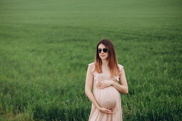 Красивая молодая беременная женщина в шляпе стоит на зеленом поле, прислонившись к стогу сена. Солнечный летний день. Она счастлива.