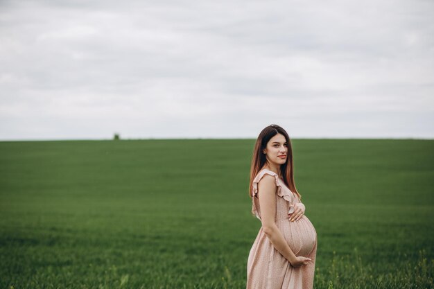 A beautiful young pregnant woman in the hat stands on a green field leaning against a haystack Sunny summer day She is happy