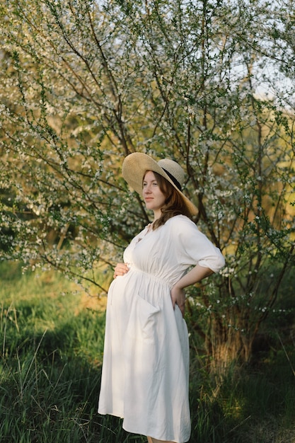 A beautiful young pregnant woman in a white dress walks in the spring garden