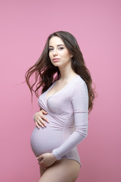 Красивая молодая беременная женщина с распущенными длинными волнистыми волосами стоит на изолированном розовом фоне и обнимает живот.