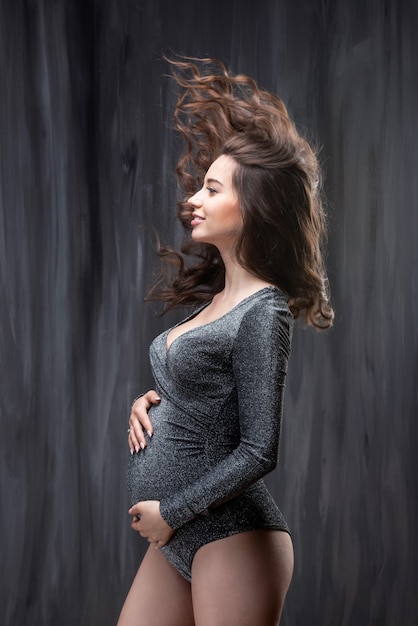 Beautiful young pregnant woman with scattering long wavy hair against a gray concrete wall. wind blowing hair.