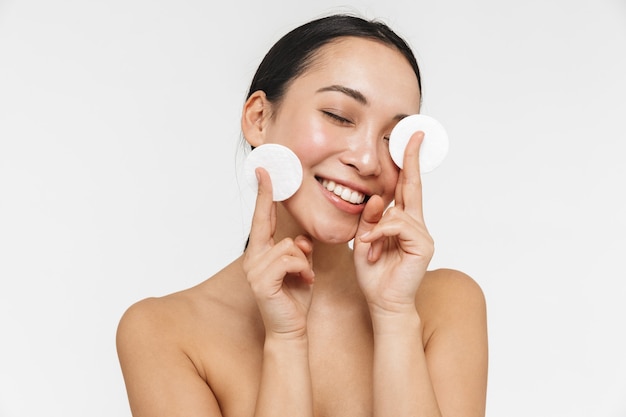 beautiful young pretty asian woman with healthy skin posing naked isolated over white wall holding cotton pads.