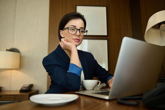 美しい若い繁栄しているビジネスマン、成功した女性のセールスマネージャーは、ラップトップで進捗レポートを書くこと、テーブルに座って、彼女のオフィスでコーヒーを飲むことに焦点を当てました
