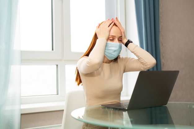 beautiful young red-haired female in medical mask at home with laptop