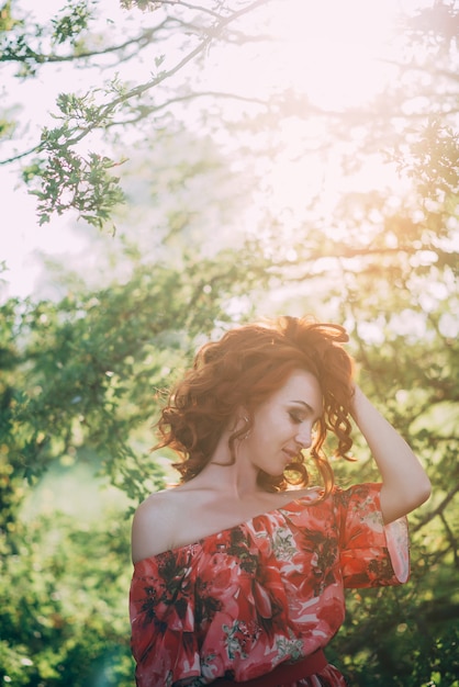 Beautiful young red-haired girl in red dress among the green foliage of tree in the backlight. 