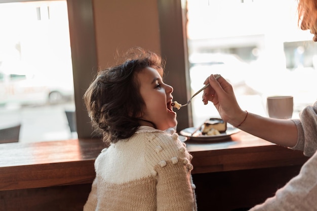 かわいい巻き毛の娘と美しい若い兄弟分母は居心地の良いカフェに座って、暖かい学校の母の日を飲んでいます。