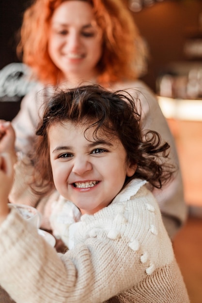 Beautiful young redhaired mother with cute curlyhaired daughter are sitting in cozy cafe and drinking hot school Mothers Day Warm