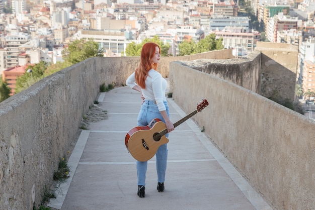 Beautiful young redhead walks with her guitar outdoors along a wall that has panoramic views of the city