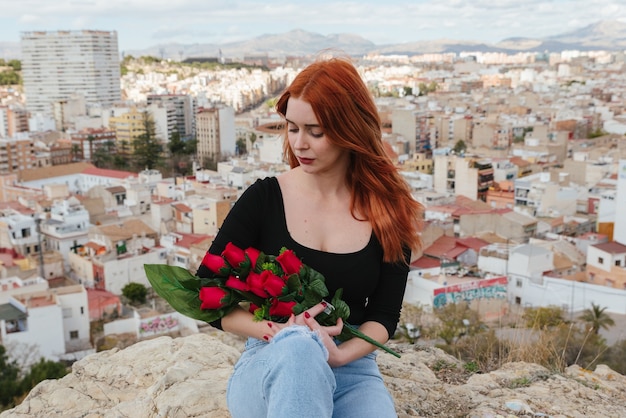 Beautiful young redhead woman in love, looks tenderly at the red roses in her arms. Valentine's Day