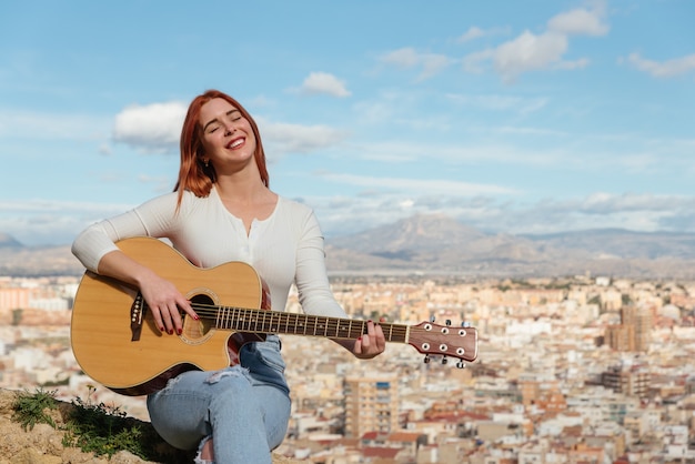 La bella giovane donna della testarossa suona la chitarra all'aperto che si siede su un banco di pietra della parete