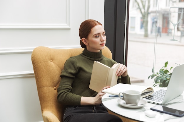 Beautiful young redhead woman relaxing at the cafe table indoors, drinking coffee, reading a book