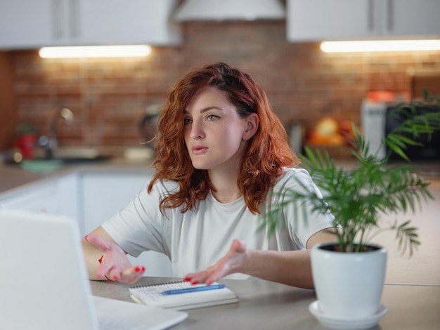 A beautiful young redheaded woman sits in the kitchen at the table with her laptop