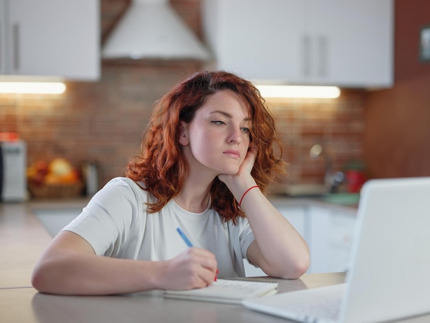 Photo a beautiful young redheaded woman sits in the kitchen at the table with her laptop