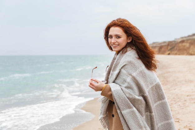 Beautiful young redheaded woman wearing coat, covered in blanket walking at the beach