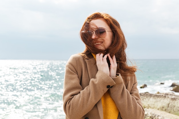 Bella giovane donna dai capelli rossi che indossa un cappotto che cammina sulla spiaggia