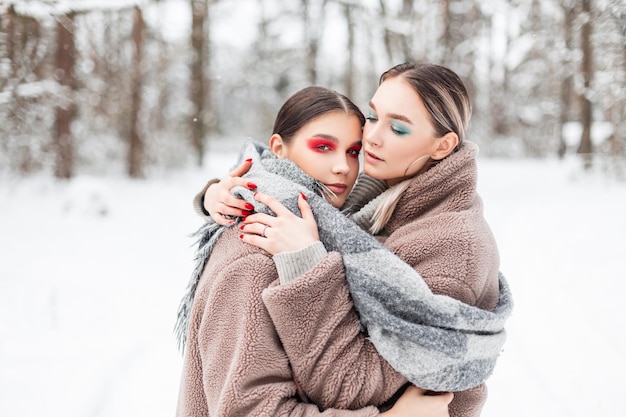 ファッショナブルな服を着た明るいメイクの美しい妹の女の子は、雪のある冬の公園で抱きしめ、暖かく保ちます