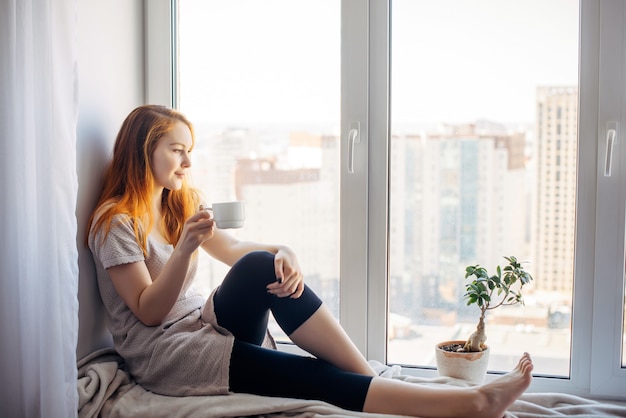 Beautiful young slender girl sitting on the windowsill at home, side view, copy space. Outside the window sky and tall city buildings. Red-haired woman drinks tea or coffee, looking at the metropolis.