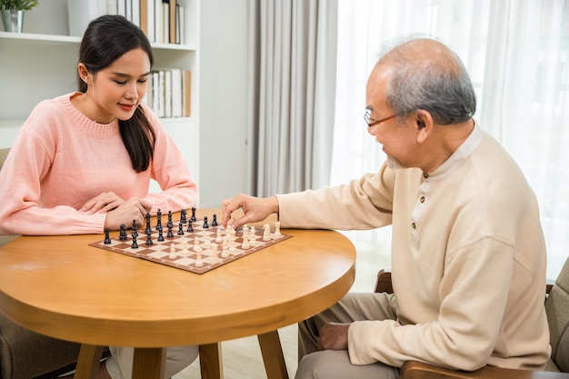 Beautiful young smile woman having fun sitting playing chess game with senior elderly at home