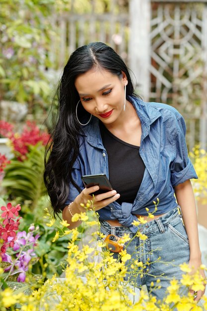 Beautiful young smiling Asian woman photographing plants and flowers in nursery for online shop or social media