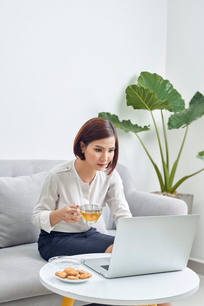 Bella giovane donna asiatica sorridente che lavora al computer portatile e che beve caffè in salotto a casa.