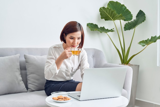 Beautiful young smiling asian woman working on laptop and drinking coffee in living room at home.