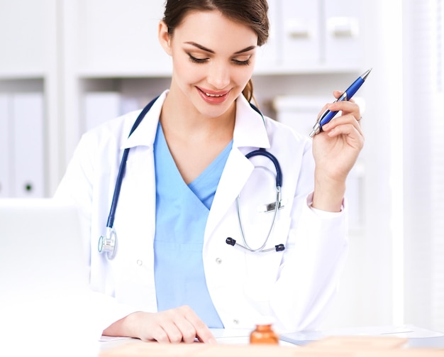 Beautiful young smiling female doctor sitting at the desk and\
writing