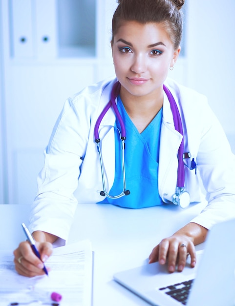 Beautiful young smiling female doctor sitting at the desk and\
writing