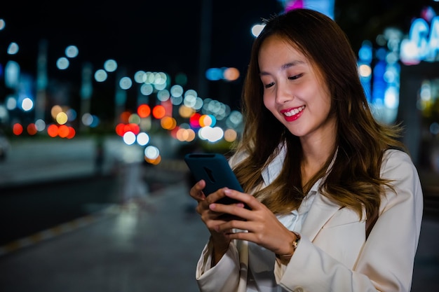 Beautiful young smiling female texting work message on smartphone outside office, Business woman using mobile phone walking through night city street while waiting car to pick up home, social media