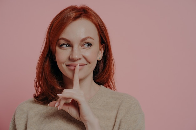 Beautiful young smiling ginger woman putting finger on lips and making hush sign