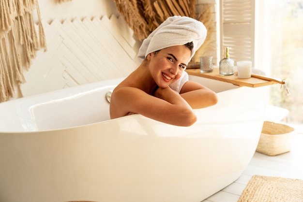 Beautiful young smiling girl relaxing lying in the bathtube in the bathroom. On her head white towel. Concept of the care and beauty at home