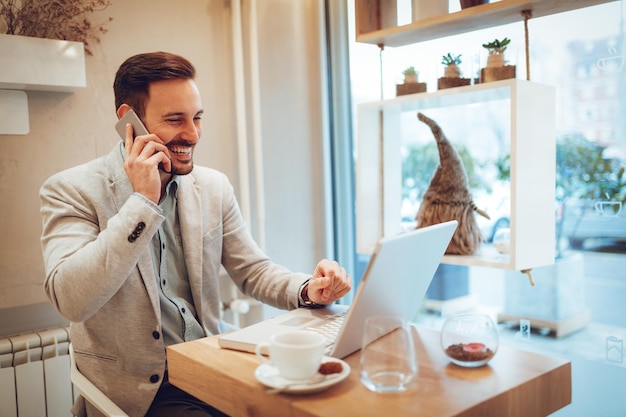 Beautiful young smiling man using smartphone and working at laptop on coffee break in a cafe.