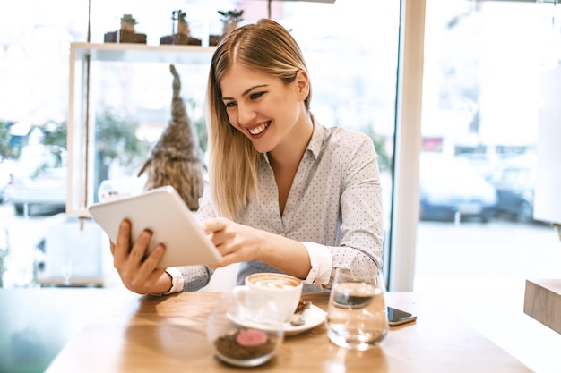 Beautiful young smiling woman drinking coffee in a cafe. She is surfing the internet on a digital tablet.