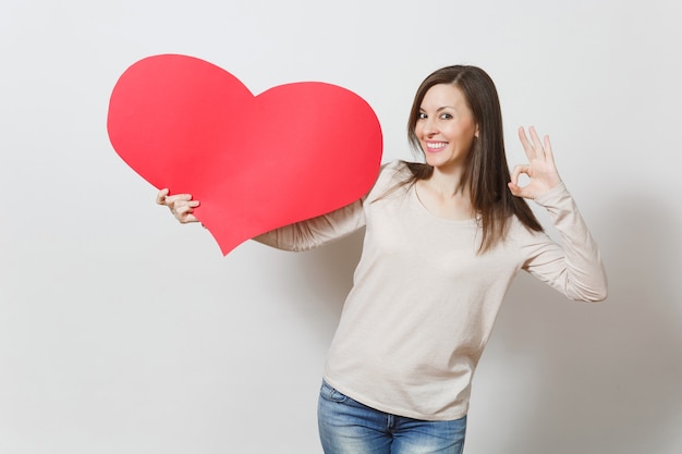 Beautiful young smiling woman holding big red heart, showing OK gesture on white background. Copy space for advertisement. With place for text. St. Valentine's Day or International Women's Day concept