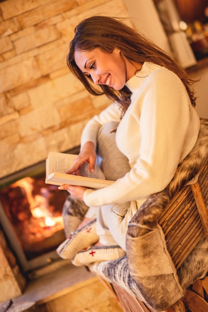 Beautiful young smiling woman reading book and enjoying by the fireplace