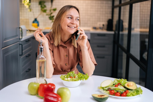Фото Красивая молодая улыбающаяся женщина разговаривает по телефону за столом с овощами, фруктами и салатом на кухне с современным интерьером. концепция здорового питания.