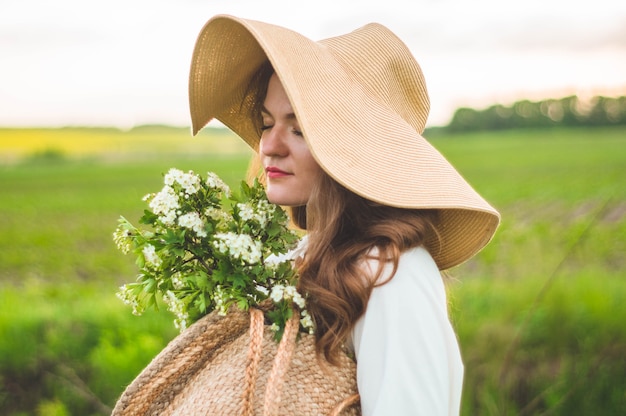 Beautiful young smiling woman in vintage dress and straw hat in field wildflowers. The girl is holding a basket with flowers