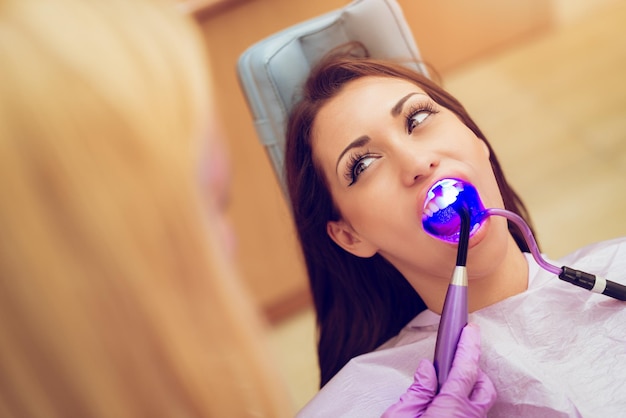 Beautiful young smiling woman at visit in the dentist office. She is sitting on a chair and dentist finishing repair tooth with dental curing UV lamp. Selective focus.