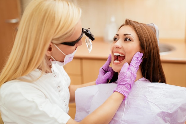 Beautiful young smiling woman at visit in the dentist office. She is sitting on a chair and female dentist checkup teeth on her. Selective focus.